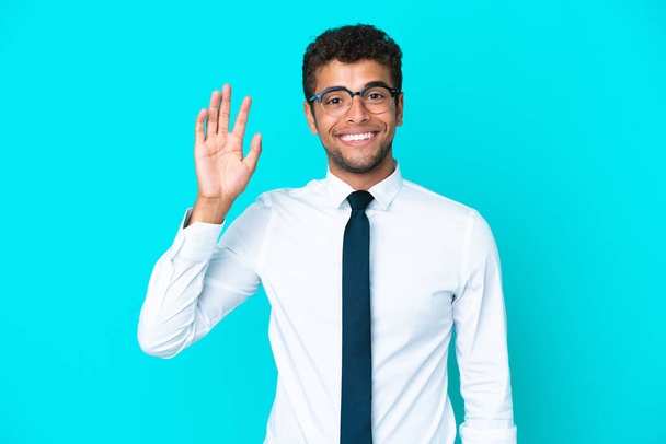 Joven hombre brasileño de negocios aislado sobre fondo azul saludando con la mano con expresión feliz - Foto, Imagen