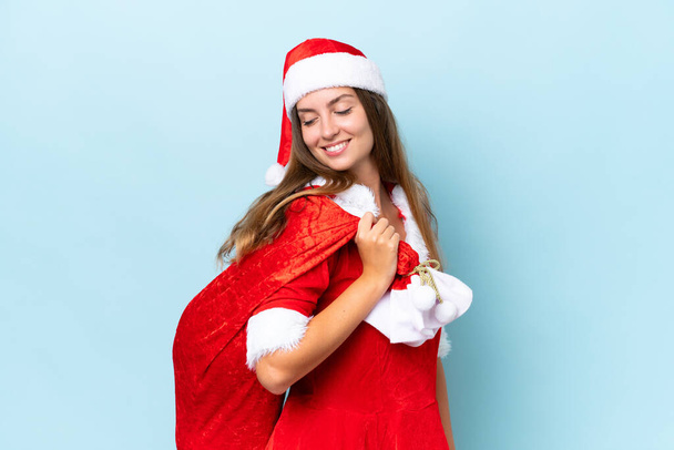 Anne Noel gibi giyinmiş genç beyaz kadın elinde Noel çuvalı tutarken mutlu bir ifadeyle mavi arka planda izole edilmiş. - Fotoğraf, Görsel