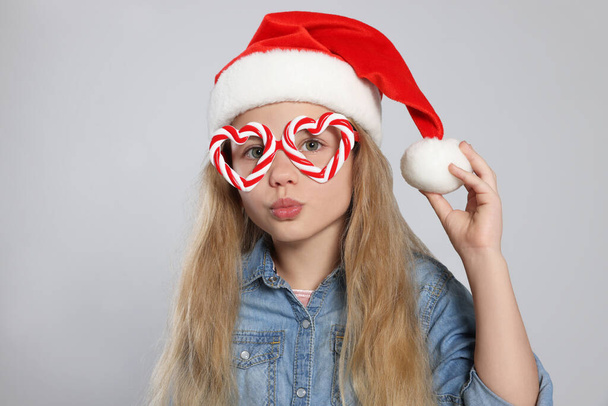 Girl wearing decorative Christmas eyeglasses in shape of hearts and Santa hat on light grey background - Photo, Image