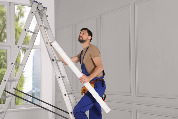 Electrician in uniform with ceiling lamp on metal ladder indoors - Foto, afbeelding