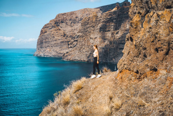 Independent relaxed calm slim girl tourist stands on the edge of a cliff with amazing ocean and sky view. Los Gigantes mountain ranges. Tenerife. Santiago del Teide. Canary Islands, Spain - 写真・画像