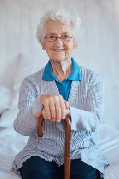 Mujer anciana, sentada retrato y sonrisa en la cama con felicidad, bastón y relajarse en el hogar de ancianos. Feliz señora mayor, dormitorio y bastón de apoyo, atención médica y bienestar en la casa de retiro. - Foto, Imagen