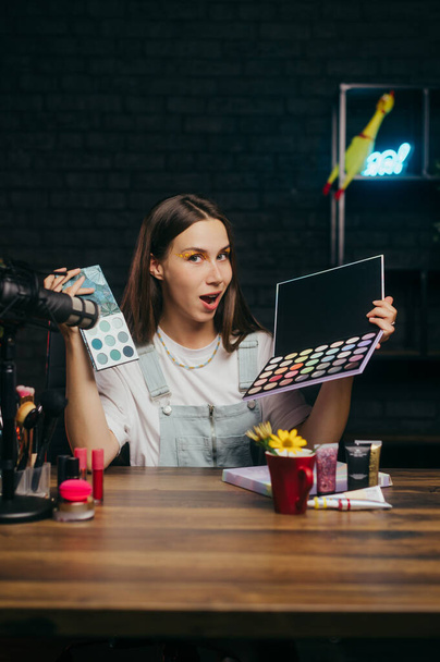 Emotional woman beauty blogger with eyeshadow palettes in hands sits at the table and records blog videos doing tests, looks at the camera with a surprised face. - Photo, Image