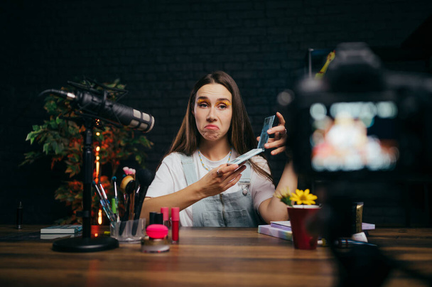 An emotional woman tests cosmetics on camera, holds an eyeshadow palette in her hands and speaks into a microphone while recording beauty videos. - Photo, Image