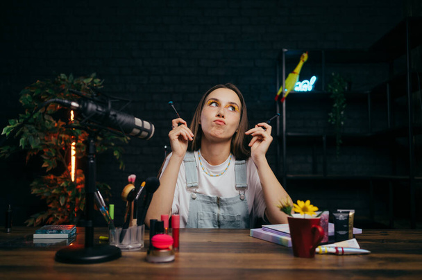 Жінка-блогер сидить з мікрофоном в студії і косметикою в руках і сумний через невдачу в записі відео, дивлячись вгору нещасно
 - Фото, зображення