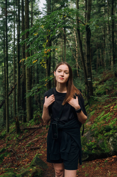 Retrato de una atractiva excursionista vestida con ropa casual parada en un sendero en el bosque durante una caminata por las montañas y mirando a la cámara con una sonrisa en la cara. - Foto, Imagen