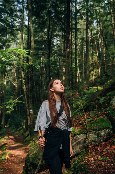 Beautiful young female tourist with a stick in her hands walks along a forest path in the mountains and looks up at the light that breaks through the trees. - Fotó, kép