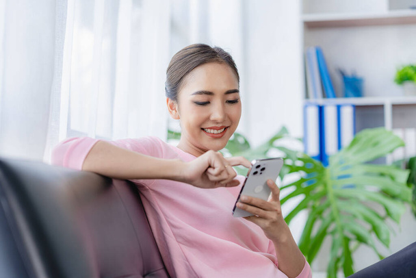 Fiatal gyönyörű ázsiai portré okostelefonnal a kanapén a nappaliban töltött szabadidő beltérben. Gyönyörű fiatal nő érzés boldog és nyugodt otthon sms chat barátaival okostelefon. - Fotó, kép