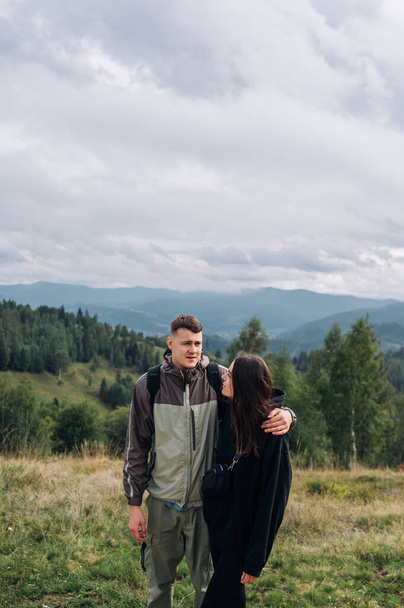 Young beautiful couple, man and woman, stand in the mountains against the background of beautiful scenery during a hike and embrace each other. Vertical photo - Фото, изображение