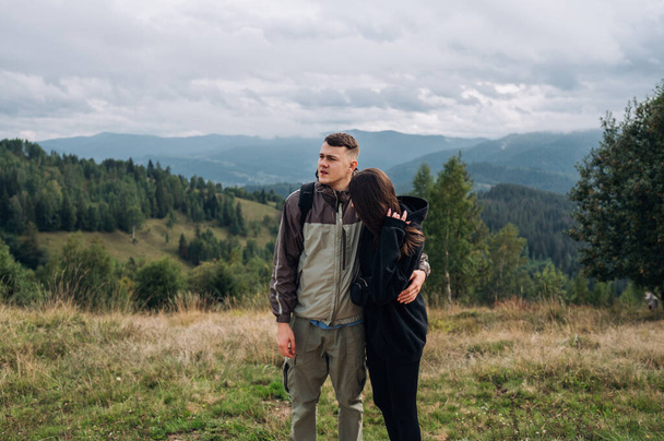 ハイキング中に山の中に立ち、美しい景色を眺めながら抱き合っている美しいカップル. - 写真・画像