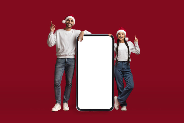 Weihnachtsliste. Glückliches arabisches Paar, das Weihnachtsmützen trägt, neben großen leeren Smartphones steht und mit erhobenen Fingern zeigt, fröhliche Ehepartner aus dem Nahen Osten, die mobile App für Online-Einkäufe empfehlen, Attrappe - Foto, Bild