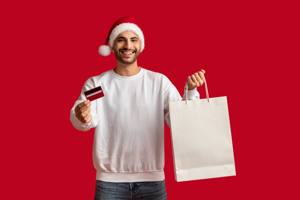 Kerstinkopen. Knappe Arabische man draagt Santa Hat Holding blanco papieren zak en creditcard, gelukkige Midden-Oosten man genietend van seizoensgebonden aanbiedingen en verkoop poseren op rode achtergrond, kopieerruimte - Foto, afbeelding