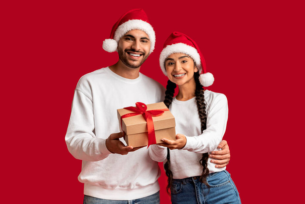 赤い背景に隔離された手の中にギフトボックスでサンタの帽子を身に着けている幸せな若いアラブカップルの肖像画,陽気中東の男性と女性はクリスマス休暇で挨拶,スペースをコピー - 写真・画像