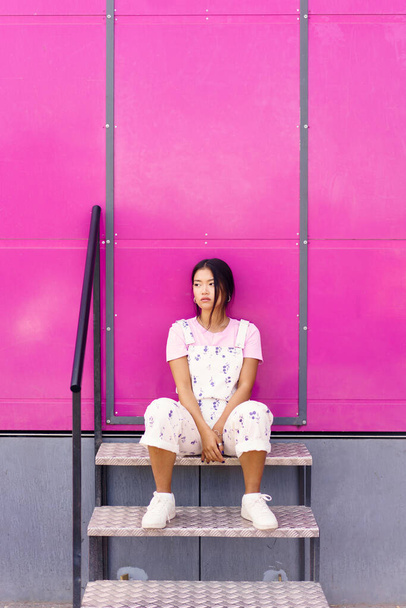 フルボディ静かな若いアジアの女性スタイリッシュな服でピンクの壁に対して金属の階段に座って離れて見て - 写真・画像
