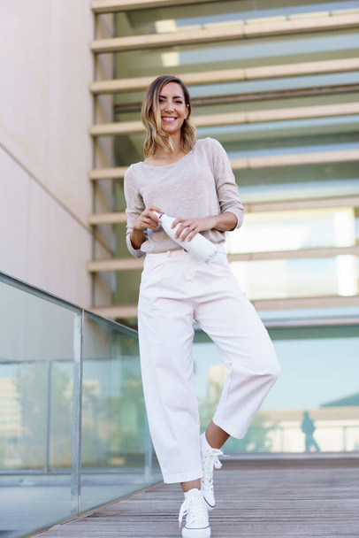Full body happy female in stylish clothes, opening bottle of water and looking at camera with smile while standing on bridge with glass railing outside contemporary building - Photo, Image