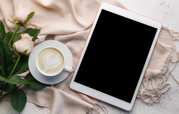 Espace de travail minimaliste avec tablette numérique écran vide maquette, écharpe confortable, une tasse de café et roses fleurs sur fond de béton. Vue de dessus, plan plat - Photo, image
