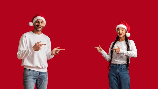 Świetna promocja. Happy Arab małżonkowie w Santa kapelusze wskazując na skopiować przestrzeń w środku stojąc izolowany nad czerwonym tle, wesoły Bliski Wschód para Wyświetlanie darmowe miejsce do reklamy - Zdjęcie, obraz