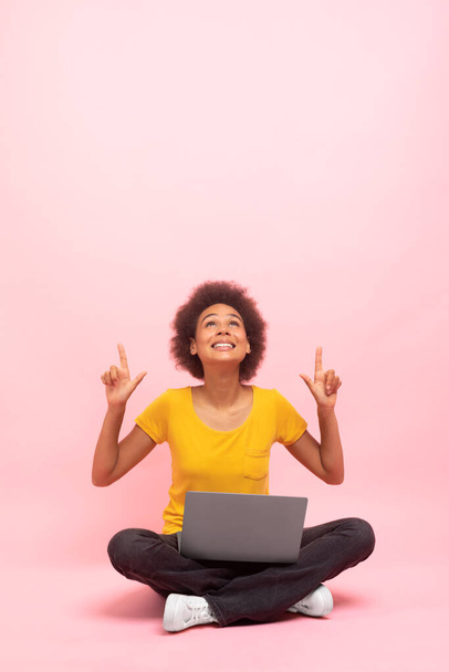Lächelnde afrikanisch-amerikanische lockige Studentin sitzt mit Laptop auf dem Boden, zeigt die Finger nach oben auf Leerraum, isoliert auf rosa Hintergrund, Studio. Schockiertes Angebot und Werbung, Gadget für Arbeit und Studium - Foto, Bild