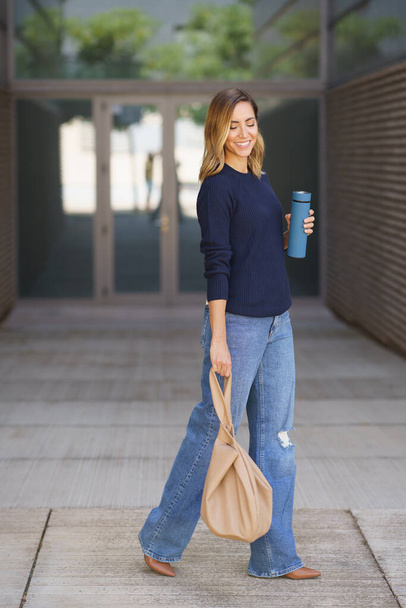 Mujer elegante positiva de cuerpo completo con termos y bolsa cerrando los ojos y sonriendo mientras camina sobre el pavimento fuera del edificio moderno - Foto, imagen