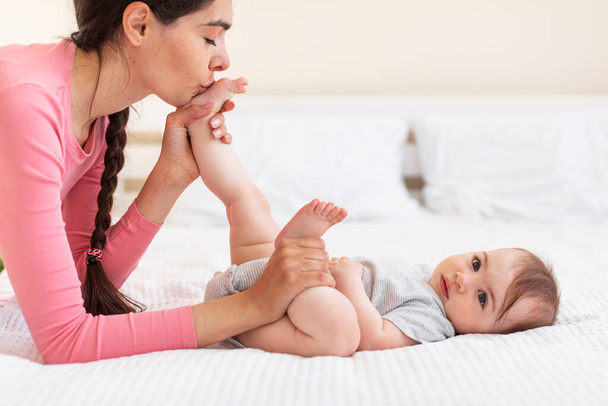 Opiekuńcza młoda matka całuje stopy niemowląt podczas gimnastyki na łóżku w domu, widok z boku, przestrzeń do kopiowania. Kochająca mama spędza czas ze swoją słodką córeczką - Zdjęcie, obraz