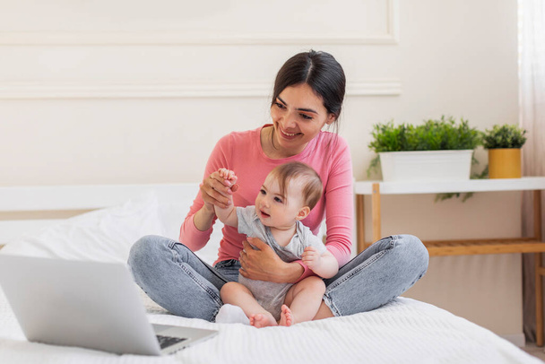 Szczęśliwa młoda matka i niemowlę dziecko za pomocą laptopa w domu, mając wideo rozmowy z rodziną, słodkie dziecko macha ręką na kamerce internetowej, siedząc razem na łóżku - Zdjęcie, obraz
