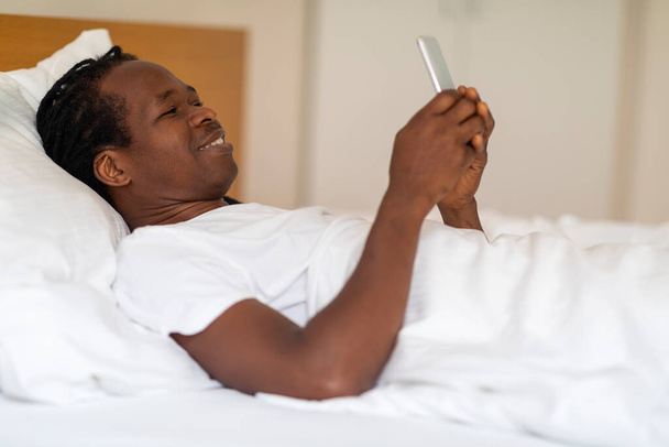 Lächelnder Schwarzer beim Surfen in einer neuen App auf dem Smartphone im Bett, glücklicher junger Afroamerikaner mit modernem Handy beim Entspannen im gemütlichen Schlafzimmer zu Hause, Seitenansicht, Nahaufnahme - Foto, Bild