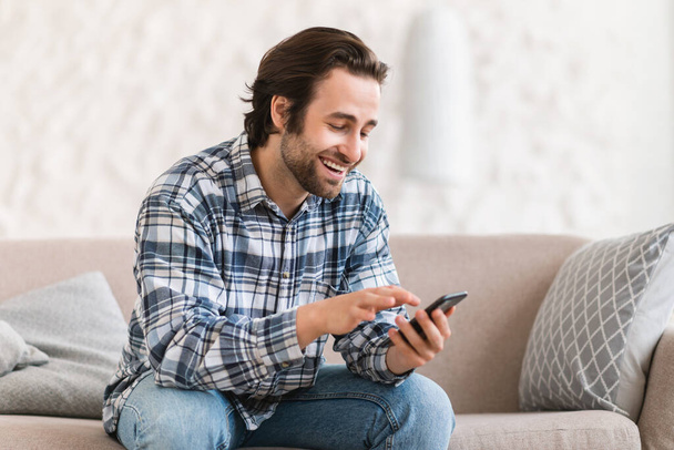 Glücklicher Millennial-Kaukasier in häuslicher Kleidung mit Stoppeln sitzt auf dem Sofa, plaudert auf dem Smartphone, schaut sich in der Freizeit Videos im Innenraum an. Neue App, Kommunikation aus der Ferne und soziale Distanz - Foto, Bild