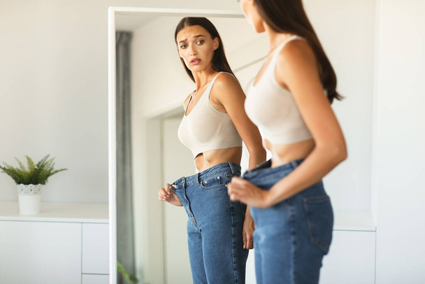 Shocked young woman wearing too big loose jeans after successful weight loss, posing and looking at her reflection in mirror with open mouth, copy space - Foto, afbeelding