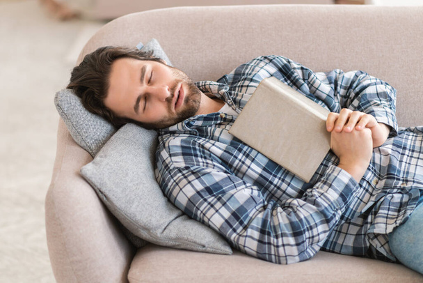 Fatigué homme millénaire européen en vêtements domestiques avec chaume dort avec livre, journal, se trouve sur le canapé dans le salon intérieur, espace de copie. Etude, éducation, surmenage, repos et détente à la maison seule - Photo, image