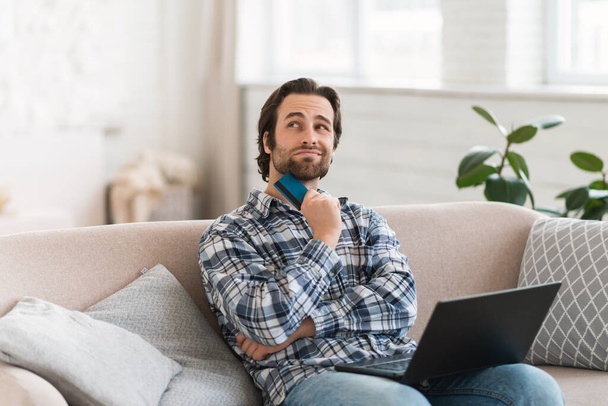 Smiling pensive millennial caucasian man in domestic clothes with stubble dreams of new buy, sit on sofa with computer and credit card shopping online in living room interior. Banking at gadget, sale - Photo, Image