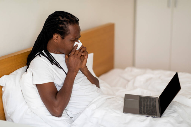 Ill Black Man φυσώντας μύτη και χρησιμοποιώντας φορητό υπολογιστή με λευκή οθόνη, ενώ κάθεται στο κρεβάτι στο σπίτι, άρρωστος αφρικανικός Αμερικανός άνδρας ελέγχοντας Flu διορθωτικά μέτρα σε απευθείας σύνδεση ή έχοντας βιντεοκλήση με το γιατρό, Mockup - Φωτογραφία, εικόνα