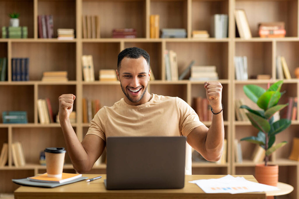 Успех бизнеса. Радостный араб, использующий ноутбук и жестикулирующий да, пожимающий кулаки, празднующий большую удачу, работающий дома, сидящий на рабочем месте - Фото, изображение