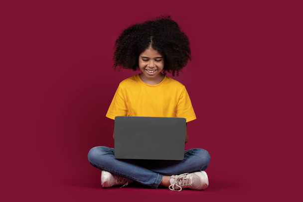 Wesoły cute czarna dziewczyna z krzaczastymi włosami w casual szkoły strój siedzi na podłodze, przy użyciu nowoczesnego laptopa, izolowane na kolorowe tło studio bordowy, przestrzeń kopiowania. Uzależnienie od dzieci i gadżetów - Zdjęcie, obraz