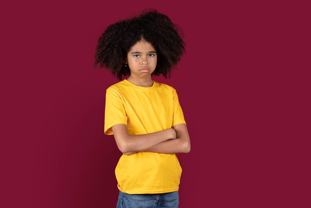 Offended black child cute girl schooler in casual outfit with bushy hair posing over colorful burgundy studio background, holding arms crossed on chest, feeling upset or displeased, copy space - Photo, Image