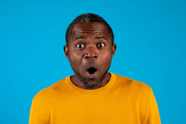 Знімок шокованого зрілого темношкірого чоловіка в жовтому, дивлячись на камеру з відкритими очима і ротом, ізольовано на синьому студійному фоні, крупним планом
 - Фото, зображення