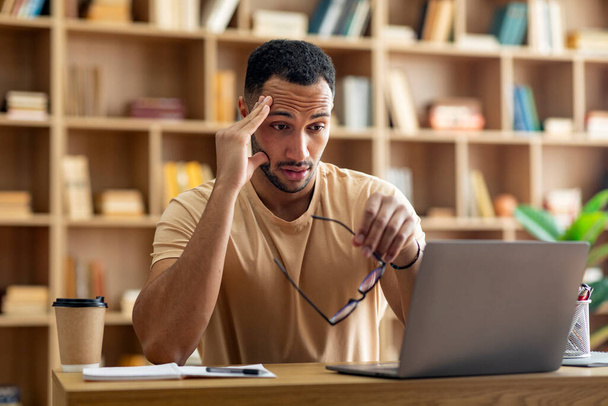 Smutný unavený arabský samec pracující na volné noze na notebooku a dotýkající se hlavy, držící brýle, dívající se na obrazovku počítače, sedící doma na pracovišti. Lhůta, přepracování, problémy při práci a stres - Fotografie, Obrázek