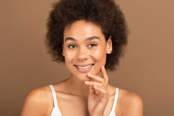 Cheerful millennial african american curly female in white top looking at camera isolated on brown background, close up, studio. Beauty and skin care at home, nude makeup, cosmetology due covid-19 - Photo, Image