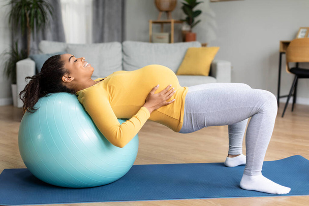 Šťastná mladá černá těhotná dáma s velkým břichem dělá cvičení na fitness míč, cítí miminko pohyby v obývacím pokoji interiéru. Sport, cvičení, tělesná a zdravotní péče doma sám a očekává dítě - Fotografie, Obrázek