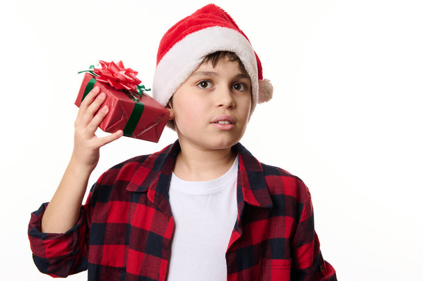 Utelias lapsi poika Santa hattu, ravistelee joululahja laatikko, arvaamalla läsnä sisällä, eristetty lähikuva muotokuva valkoisella taustalla kopioi tilaa. Aika avata lahjat. Hyvää talvilomaa - Valokuva, kuva