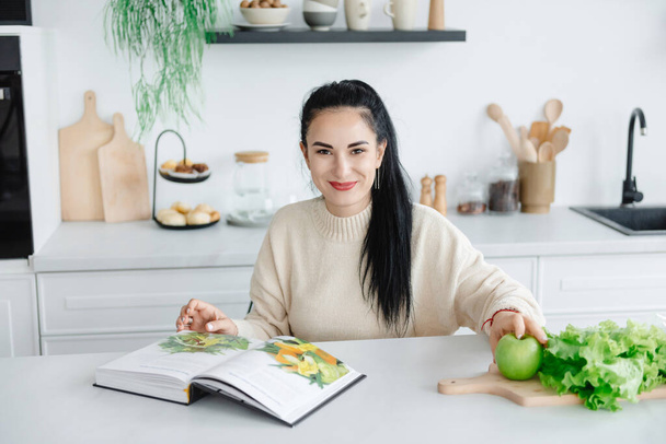 Una joven optimista mira a la cámara y sonríe mientras se sienta en su cocina. Leer una revista sobre alimentación saludable y estilo de vida. - Foto, imagen