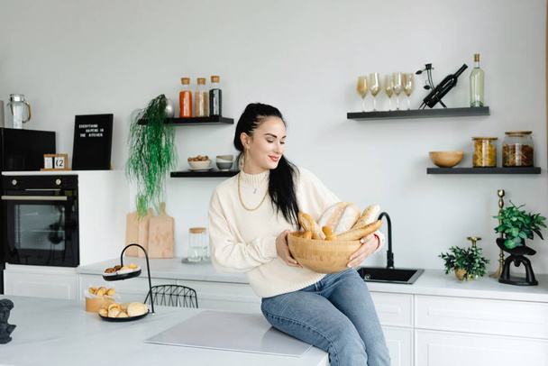 Nainen istuu keittiössään leipäkori kädessä. Nautin pikkuasioista. Lykken käsitteet. Rentoutumisen käsitteet. - Valokuva, kuva