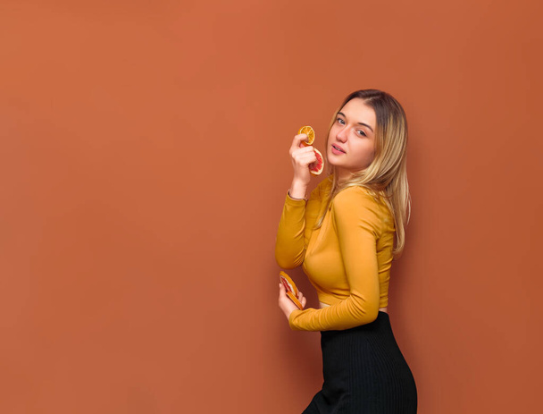 Joven chica atractiva sostiene toronja y rodajas de naranja en sus manos, mira a la cámara, de pie de lado, en blusa amarilla y fondo negro sobre fondo de vainilla. - Foto, imagen