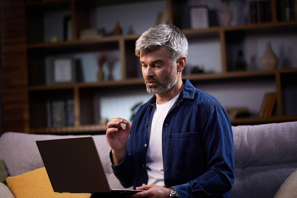 A male freelancer using a laptop works remotely and sits on the couch. Focused man participating in video conference on laptop computer while working remotely at home. - Photo, Image