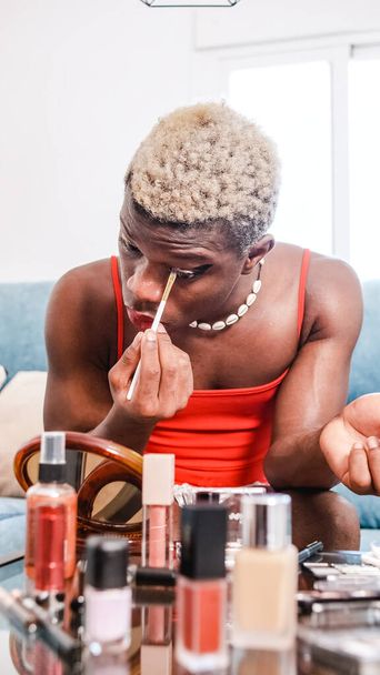 Afro-américain en robe avec collier coquillage regardant dans le miroir et appliquant fard à paupières tout en étant assis sur le canapé à la maison - Photo, image