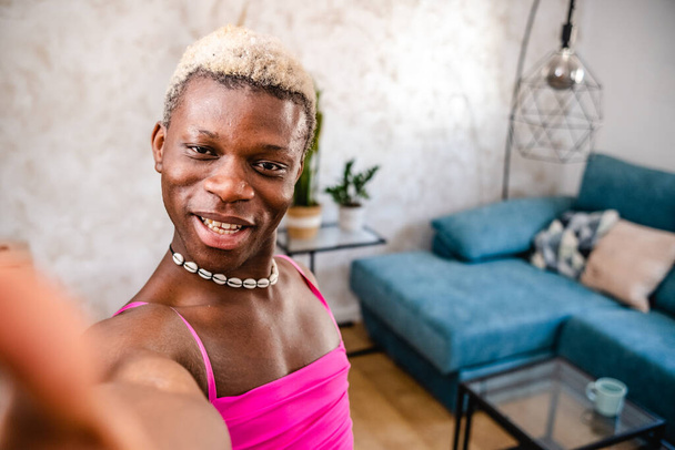 Από τα πάνω χαρούμενα νεαρή Αφροαμερικανή τρανσέξουαλ γυναίκα με βαμμένα μαλλιά σε ροζ φόρεμα χαμογελώντας έντονα και κοιτάζοντας την κάμερα, ενώ χορεύει και δείχνει το σπίτι της - Φωτογραφία, εικόνα