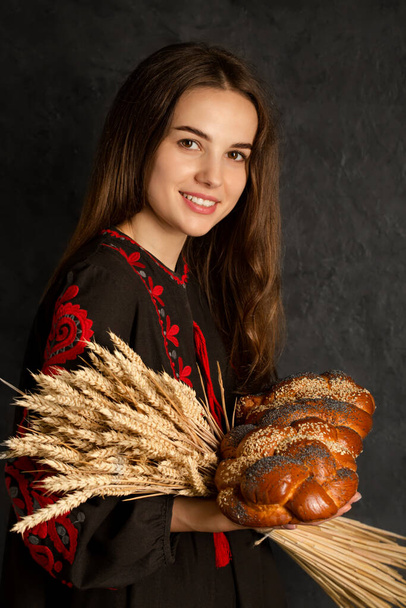 Een Oekraïense vrouw in een geborduurde jurk houdt een broodje en een boeket tarweoren vast - Foto, afbeelding