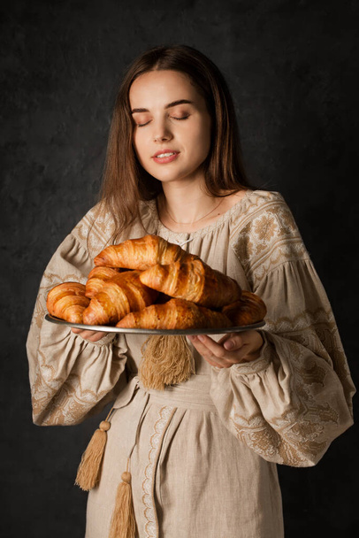 Ukrainian woman in national dress holds a tray with rosy croissants on a dark background - Foto, afbeelding
