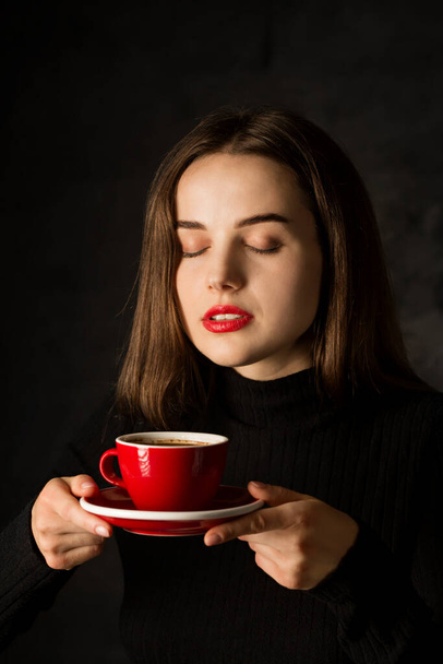 Όμορφη νεαρή γυναίκα με ένα κόκκινο φλιτζάνι ευωδιαστό καφέ σε σκούρο φόντο - Φωτογραφία, εικόνα
