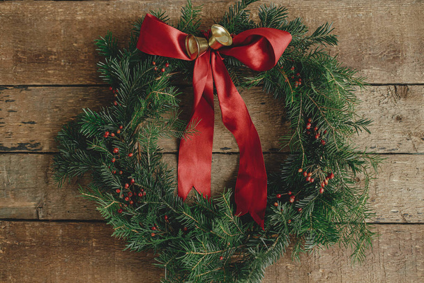 Christmas wreath flat lay.Stylish christmas wreath with red ribbon, red berries and golden bells on rustic wooden table. Winter holiday decor. Merry Christmas and Happy Holidays! - 写真・画像