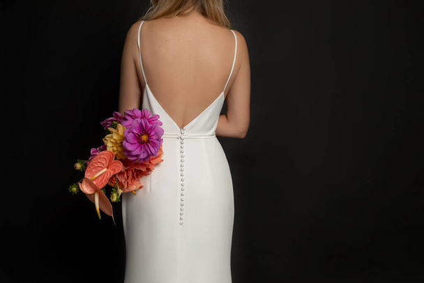 Rearview of unrecognizable, cropped blond woman with naked back in white buttoned dress hold bunch of multicolored flowers in black studio. Wedding, marriage, engagement concept. Copy space, mockup - 写真・画像